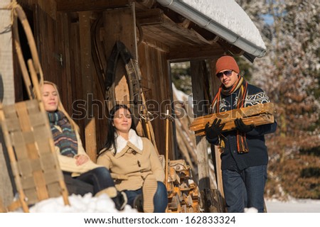 Young man carry wooden log winter cottage snow girlfriends sunbathing