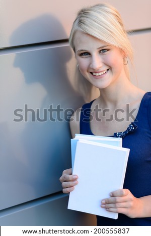 Smiling blond student girl with books outside leaning against wall