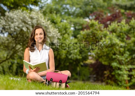 Smiling student girl sitting on grass with open book summer