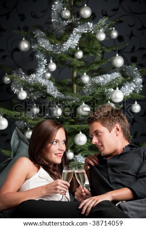 Young extravagant man and woman with champagne sitting in front of silver decorated Christmas tree