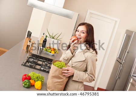 Young woman unpacking shopping bag with grocery in kitchen, biting apple