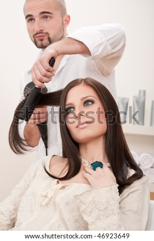 Professional male hairdresser with hair dryer and hair brush drying hair at salon with female customer