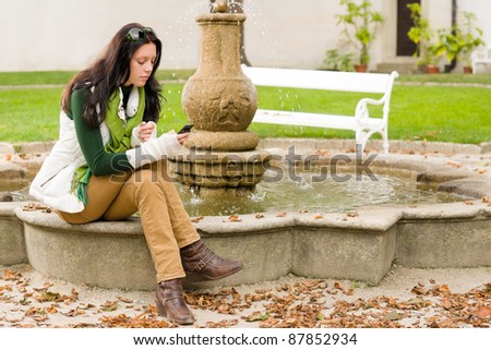 Autumn park young woman dialing phone sitting on fountain