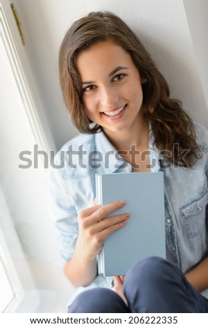 Teenage girl student sitting by window holding book smiling at camera