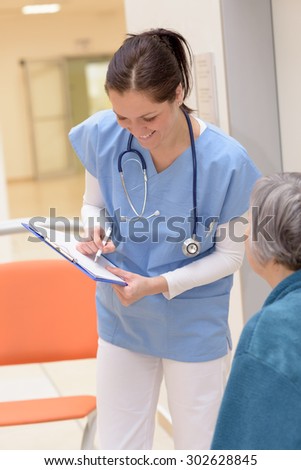 Smiling young female nurse writing in senior patient file in hospital