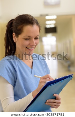 Smiling young female nurse in hospital corridor writing in patient file