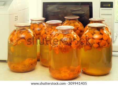 Home canning: large glass cylinders with apricot compote, sealed with metal caps. Presented on the background of a window.