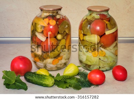 Home canning: large glass cylinders with a variety of vegetables: cabbage , tomatoes, cucumbers, peppers. Sealed with metal caps. Located next to fresh vegetables.