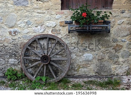 An old wooden wheel leaning against a stone wall decorated with a pot of red geraniums