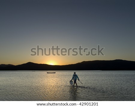 An unrecognizable fisherman walks through the shallow lake to reach his boat on the magic hour.