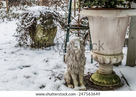 Mass production polyresin lion covered with snow besides a flower bucket with patina, concrete planter in background.\
Closeup garden with snow for book cover, flyer, magazine, CD cover design, website