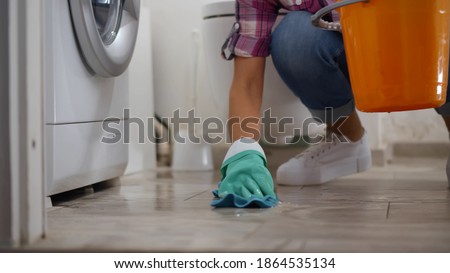 Woman in gloves having leaking washing machine wiping floor and wrinkling rag in bucket. Housewife near broken washing machine collecting water in basin in bathroom