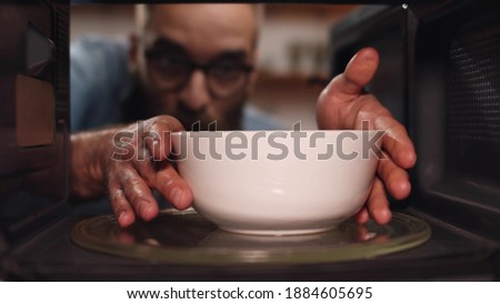 Young caucasian man heating food in microwave oven. View from inside microwave of bearded guy opening door and taking warm bowl with meal for dinner