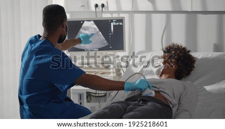 Young afro-american pregnant woman in safety at ultrasound office in modern clinic. Portrait of young male doctor wearing safety mask and gloves using ultrasound machine to check pregnant woman.