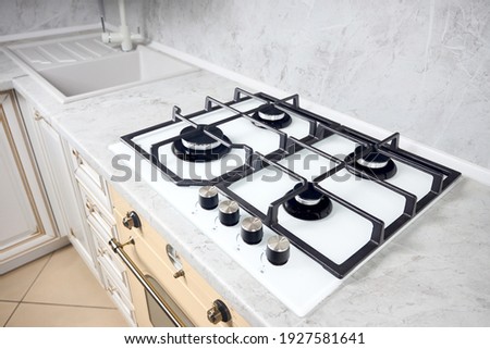 Modern white gas stove on counter top closeup. Hob gas stove made of tempered white glass using natural gas or propane for cooking on light countertop with sink faucet mixer tap in kitchen interior.