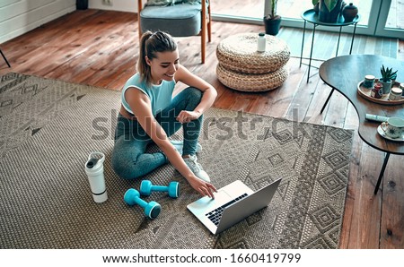 A sporty woman in sportswear is sitting on the floor with dumbbells and a protein shake or a bottle of water and is using a laptop at home in the living room. Sport and recreation concept. Imagine de stoc © 