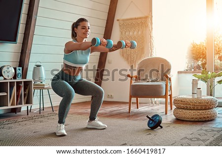 Beautiful young athletic girl in leggings and top crouches with dumbbells at home. Sport, healthy lifestyle. Stock fotó © 