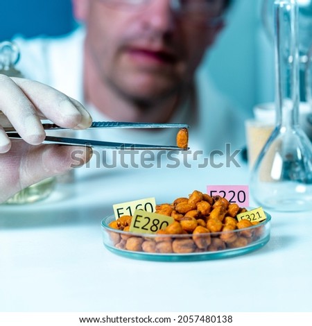 A laboratory assistant's hand holds a corn seed in tweezers, and corn seeds in a petri dish are decorated with plates with the names of E. Food Laboratory additives. 商業照片 © 