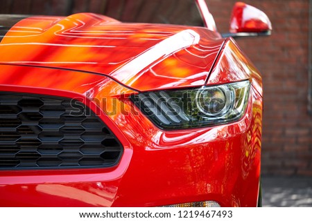 Front view of sportscar with brick wall. Concept of car detailing and paint protection background. Clos up of headlight detail of modern luxury car with reflection on red paint after wash & wax.