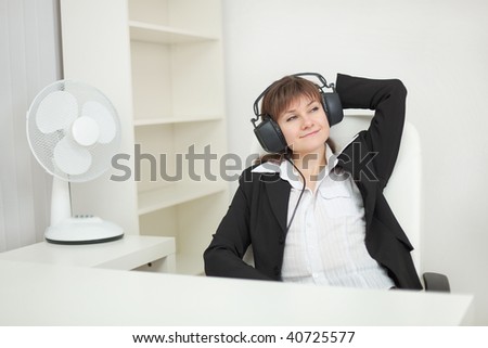 The young beautiful woman sits at office with ear-phones on a head and listens to music
