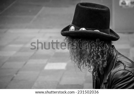 Black and white image of a person sitting in a paved urban street with long curly hair, leather jacket and hat.