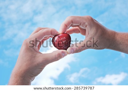 A red stone heart with I LOVE YOU carved on the front held aloft in a mans hands against a backdrop of a blue sky with clouds
