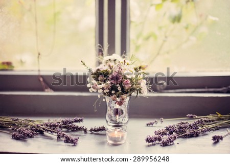 A vintage vase full of wild flowers in lilacs and creams. Vase is sat on window sill and surrounded by lavender. Vintage colour/color overlay added.