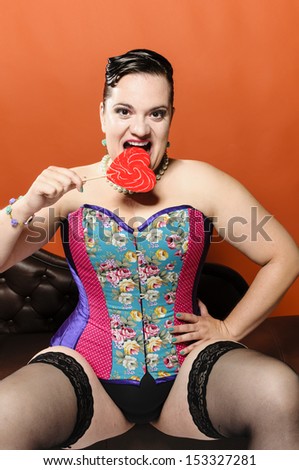 Attractive overweight woman sitting on a chaise longue and holding a big red heart shaped lollipop in hand. She wears a colorful corset, black nylon stockings. She grins and bites the lollipop.