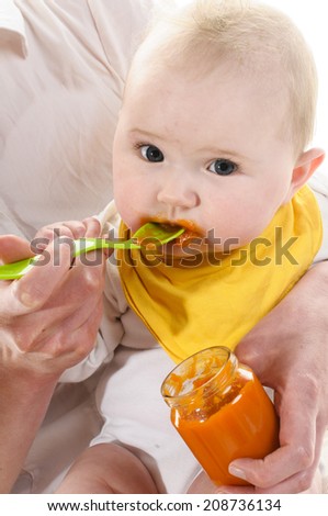 Adult woman sitting on a white chair and holds the 6 months old baby in her arms and lined with a yellow plastic spoon an orange carrot, isolated against white background.