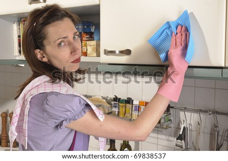 Adult housewife, cleaning lady is in her bright kitchen and cleaning with rubber gloves and rags, looking sadly into the camera.