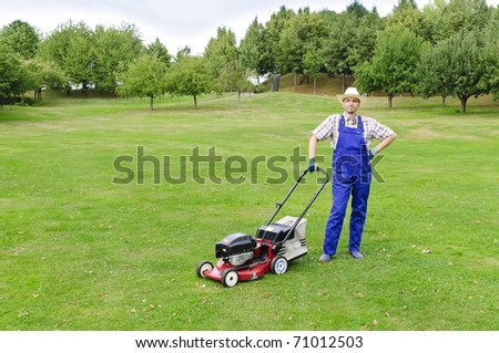 Adult man (gardener) in the blue overalls and straw hat with the lawn mower on a large lot