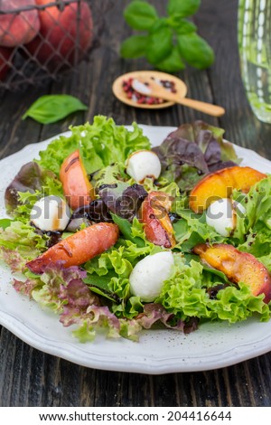 Close-up of assorted salad leaves, grilled peaches and mozzarella salad on a white vintage plate placed on a dark wooden surface; glass of water and fresh basil on the background