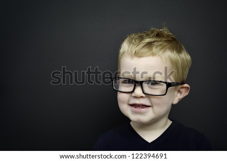 Smart young boy wearing black rimmed glasses stood smiling in front of a blackboard