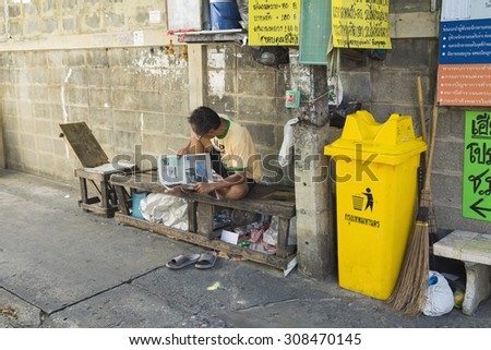Bangkok, Thailand - June 29, 2015: Old man reading newspaper on old wooden bench, next to rubbish bin, on small alley in Bangkok
