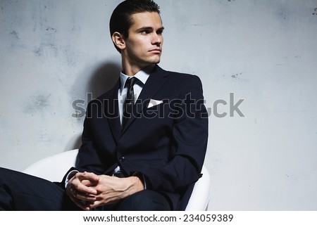 Serious man in a black business suit sitting on a white chair on a white grungy background. Studio shoot