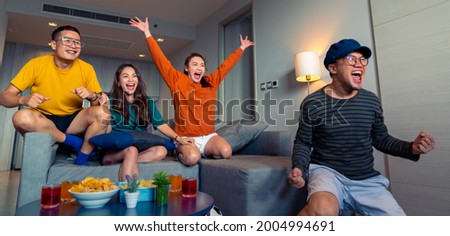 Group of Asian people friends sit on sofa watching and cheering sports games competition on TV together at home. Excited man and woman sport fans celebrate sport team victory in national sports match