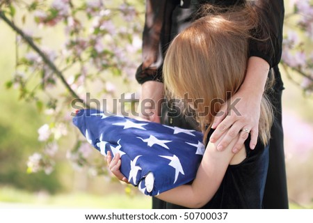 Daughter holding a parent\'s folded American flag with a woman\'s arms wrapped around her.