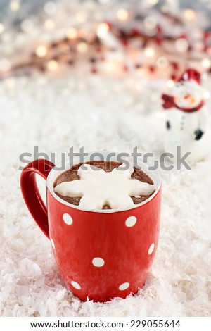 Vibrant red cup of hot chocolate with snow flake shape of whipped cream. Snowman and Christmas lights in the background. Extreme shallow depth of field.