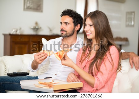 Happy couple watching tv while eating pizza. Shallow depth of field, focus on the man