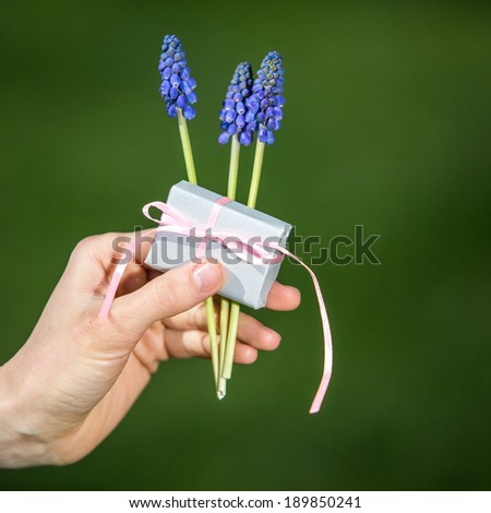 Woman holds small present gift with spring flowers in a hand over green nature background