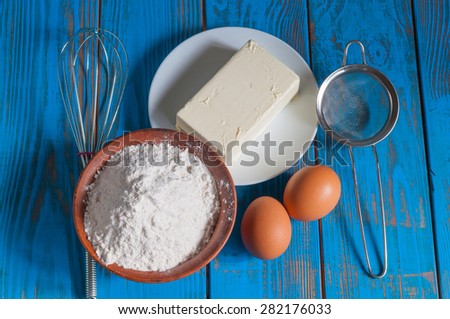 Baking cake in rural kitchen - dough recipe ingredients eggs, flour, butter, sugar and whisk, screen on vintage blue wood table from above.