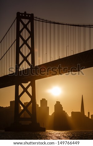 Sunset silhouettes San Francisco buildings as sailboat glides under Bay Bridge