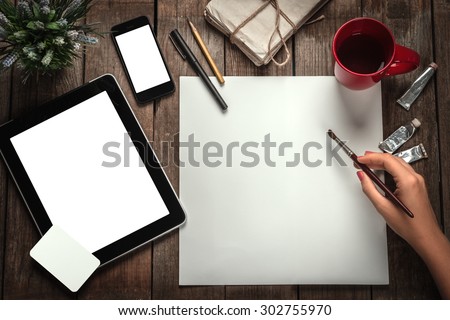 Woman\'s Hand With A Brush Paints On A Paper. Tablet Computer, Phone And Business Cards On A Wooden Table