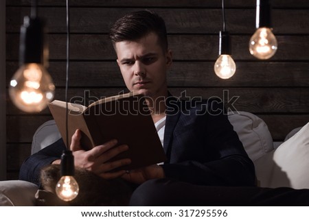 Thoughtful Handsome Man In Suit With Book Sitting On Sofa Between Lamp On Wooden Background
