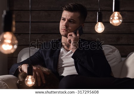 Thoughtful Handsome Man In Suit With Smartphone Sitting On Sofa Between Lamp On Wooden Background