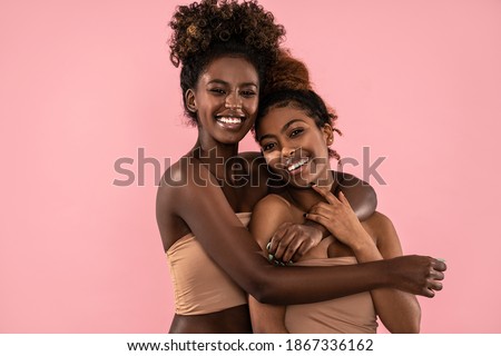 Excited emotional young women posing together , smiling and looking at camera. Pink pastel studio background. Beauty portrait of two afro female models. Perfect toothy smiles.