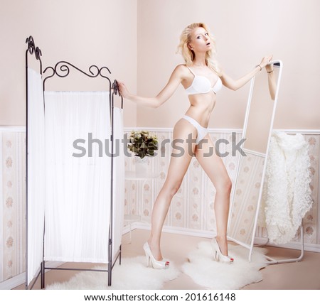 Delicate beautiful slim blonde woman posing in sensual lingerie, standing in room. Girl looking at camera