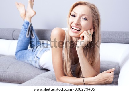 Beautiful smiling young blonde woman lying on the couch, relaxing at home. Girl looking at camera.