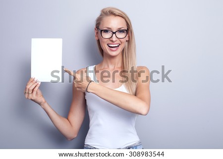 Smiling young blonde woman holding white empty board. Girl wearing fashionable eyeglasses. Studio shot.