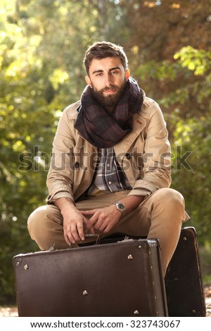 Fashion photo of young handsome man with suitcases in park. Autumn. Men wearing coat and scarf. Sunny day.